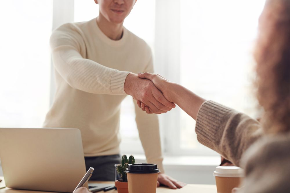 A handshake across a desk with a laptop and coffee cups